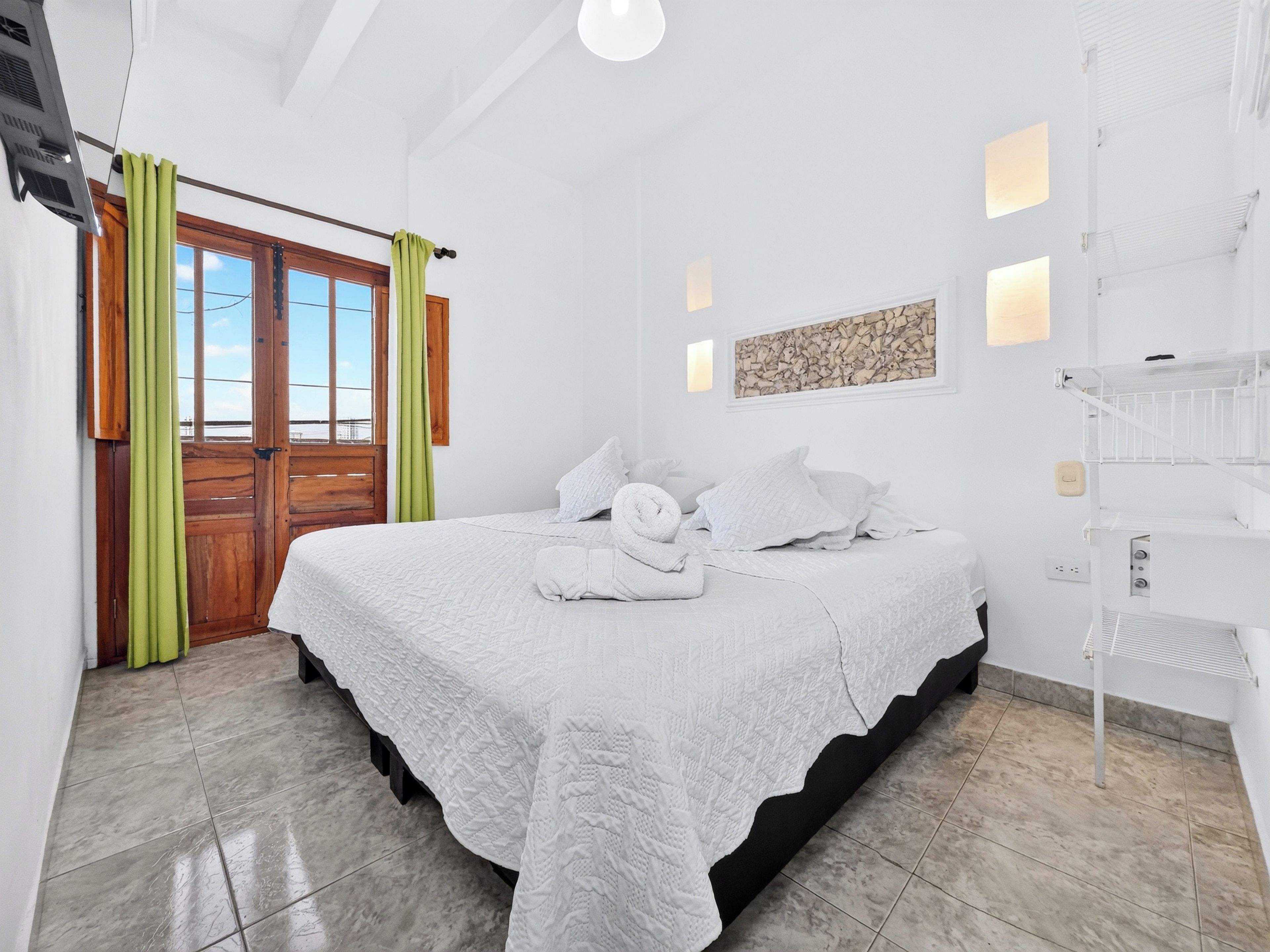 Un dormitorio con una cama, una puerta de madera, una ventana y una lámpara fijada a la pared.