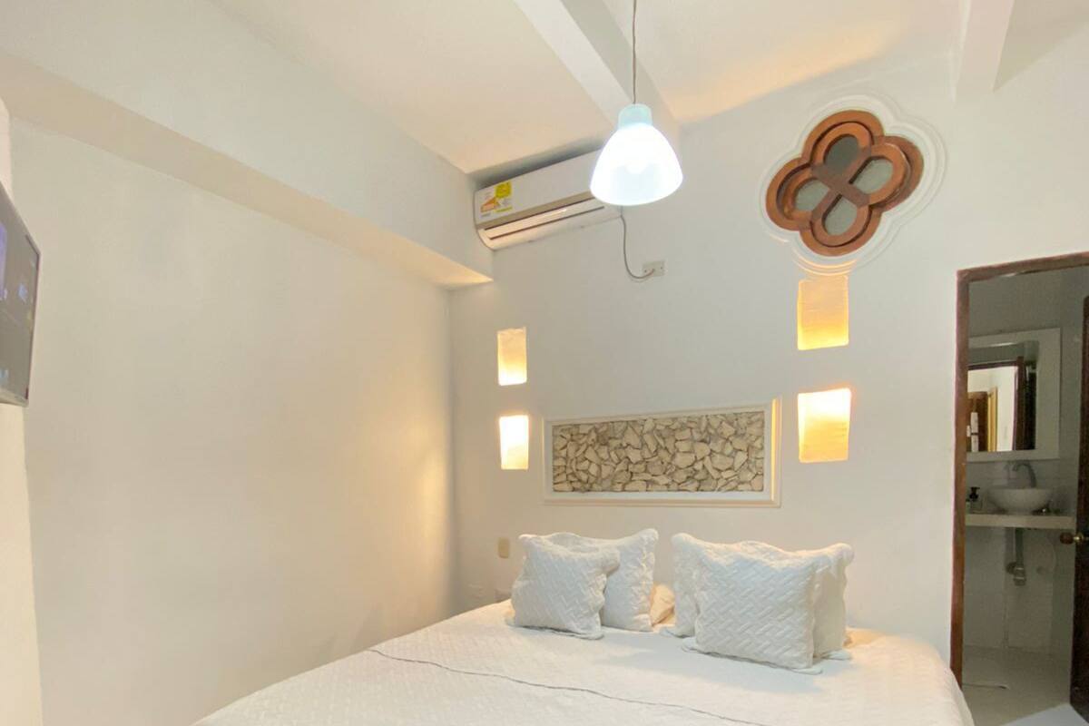 Una cama bien hecha con sábanas y almohadas blancas, una lámpara colgante y un adorno de pared decorativo.