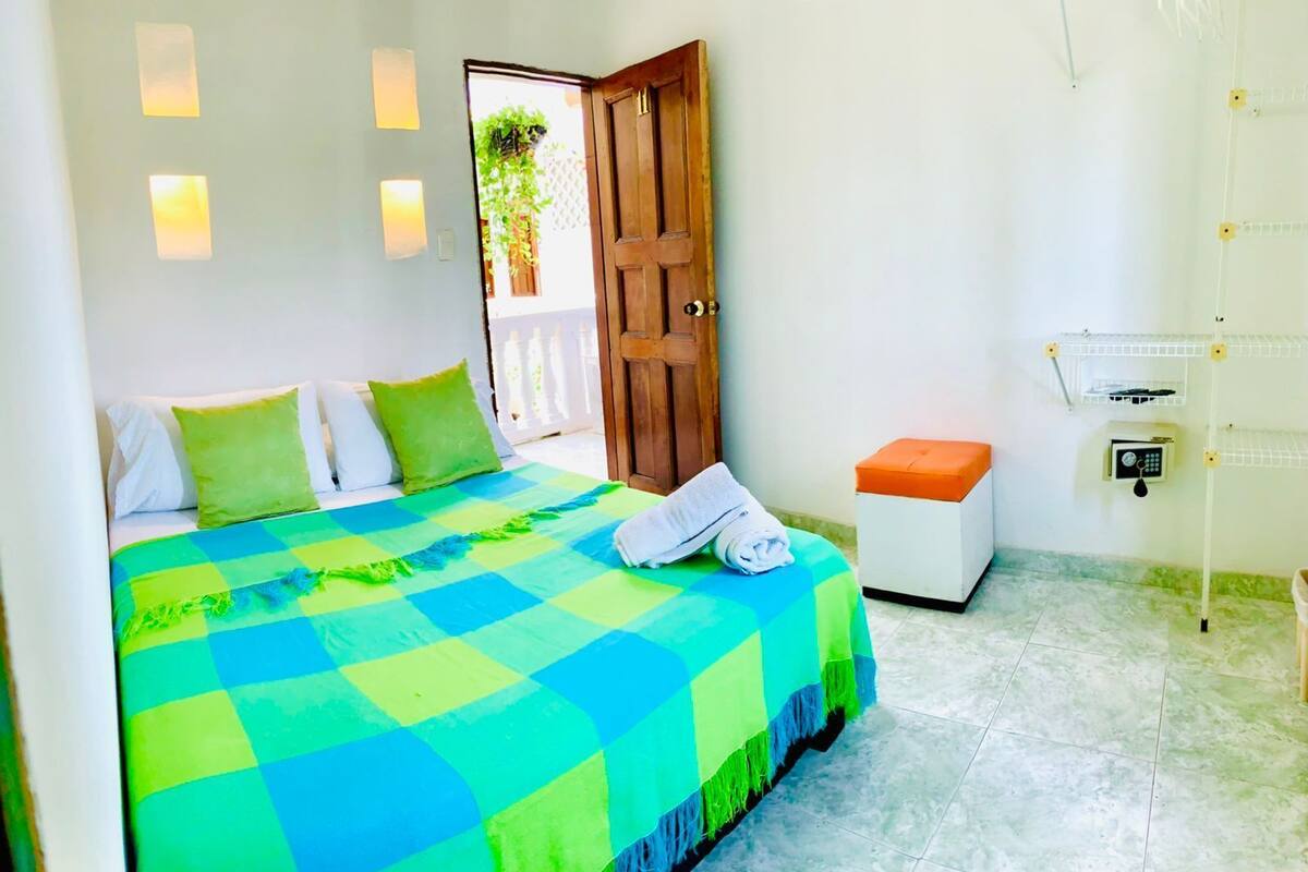 Un dormitorio con cama, almohadas, una mesita de noche, un taburete naranja pequeño, un dispositivo de seguridad fijado a la pared y una puerta.