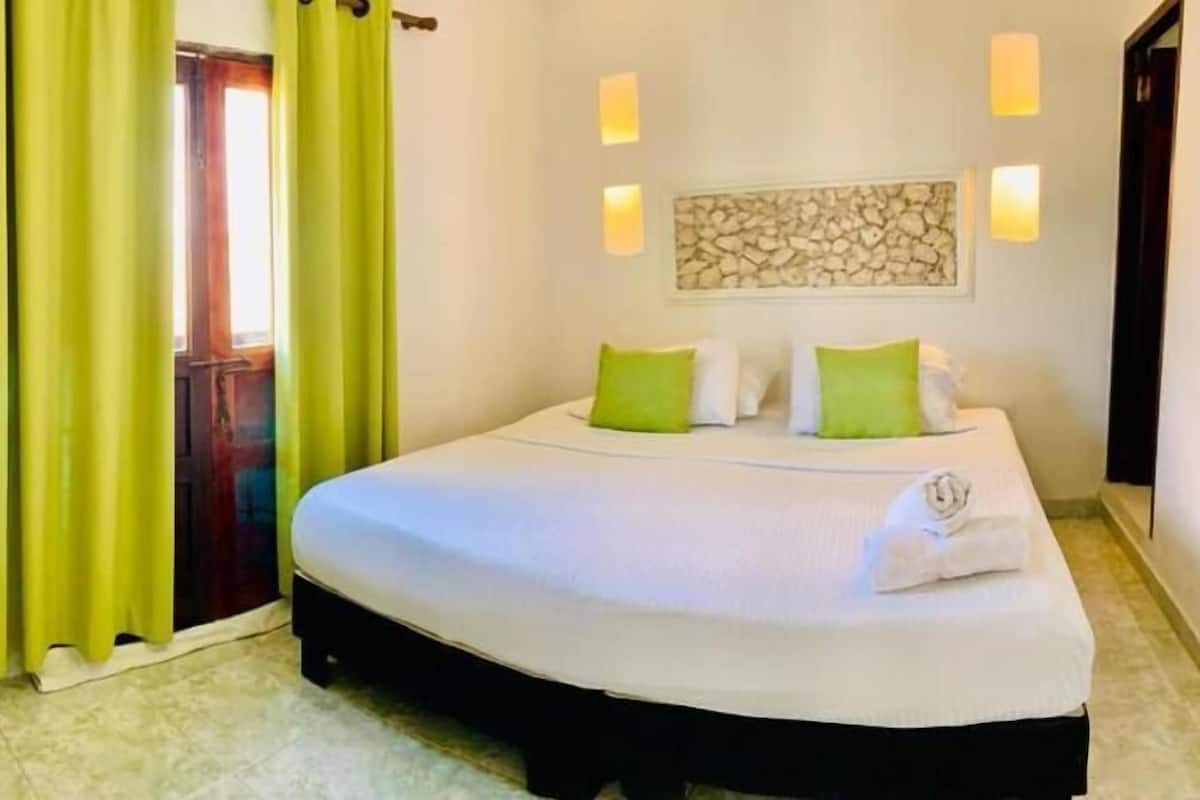 Un dormitorio con cama, almohadas verdes, una obra de arte colgada en la pared y una puerta con cortinas.