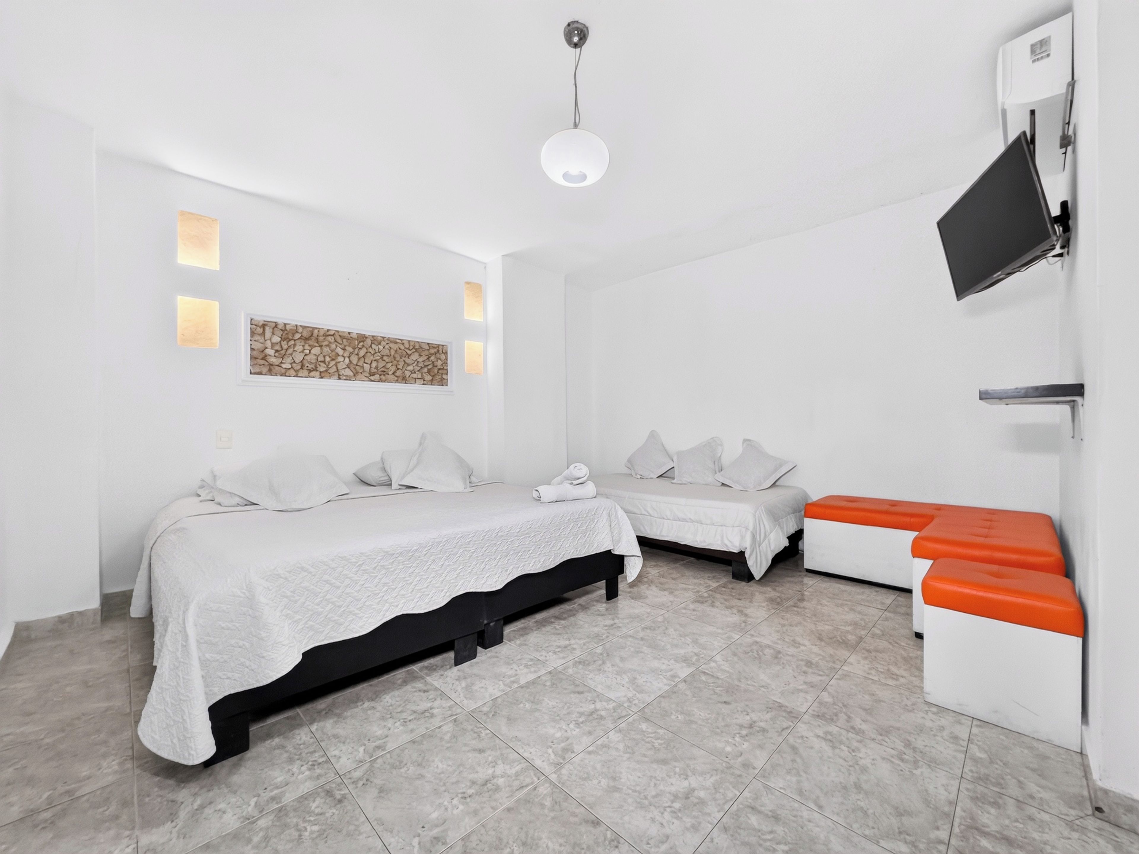 A modern hotel room with two beds, a flat-screen TV, and a seating area with orange cushions.
