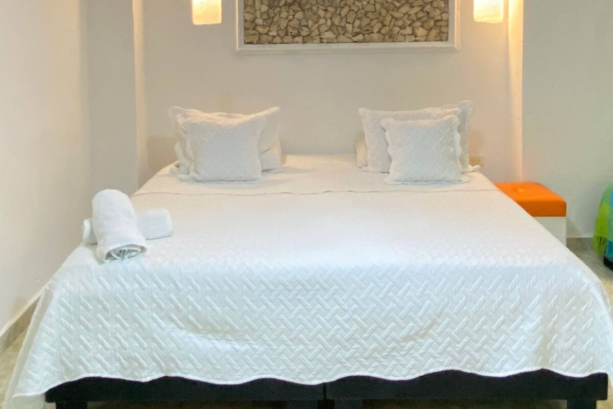 Una cama bien tendida con ropa blanca y almohadas, una obra de arte colgada en la pared y dos apliques.