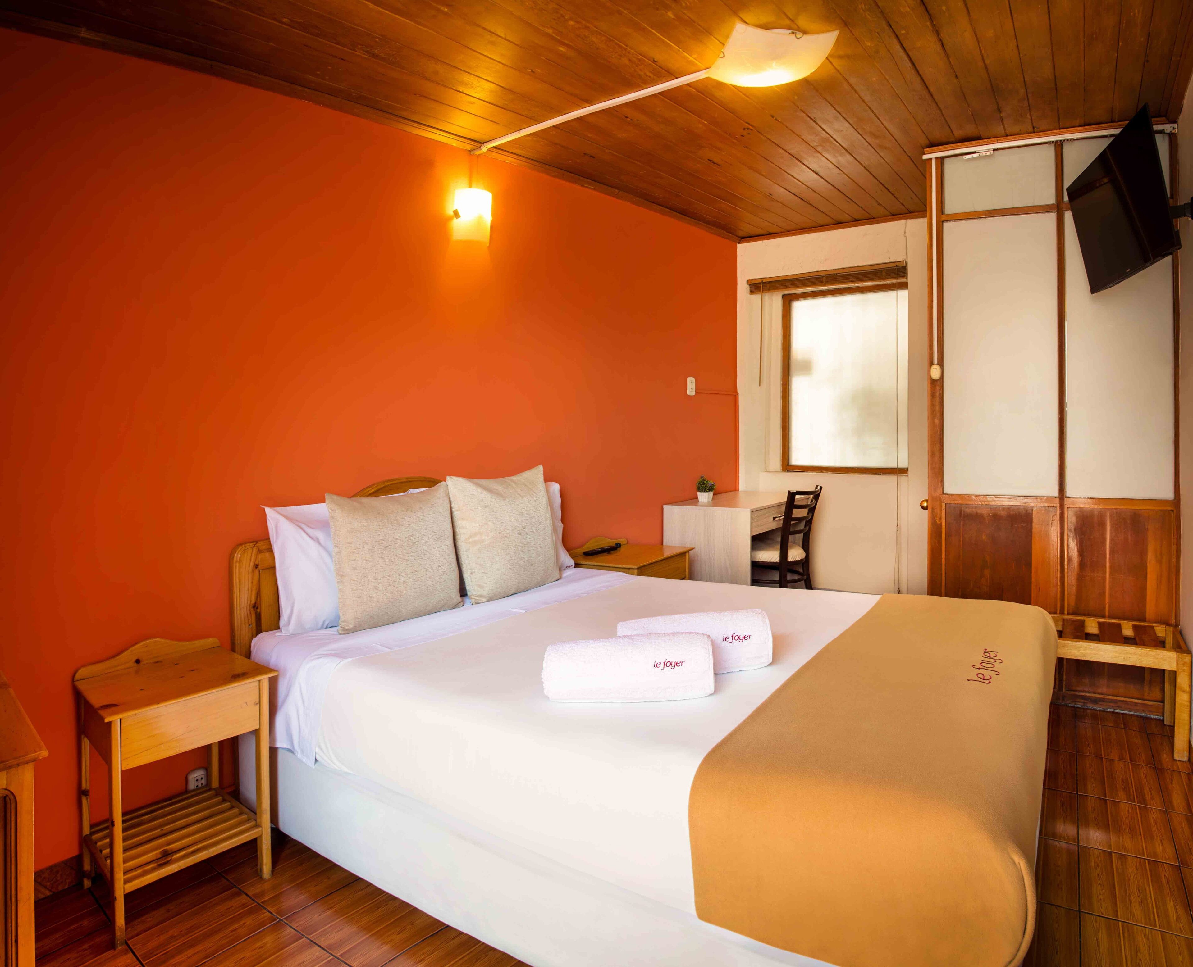 Habitación de hotel con techo de madera, cama con ropa blanca, mesita de noche, una silla, escritorio, planta pequeña en maceta y televisor montado en la pared.
