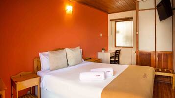 Habitación de hotel con techo de madera, cama con ropa blanca, mesita de noche, una silla, escritorio, planta pequeña en maceta y televisor montado en la pared.