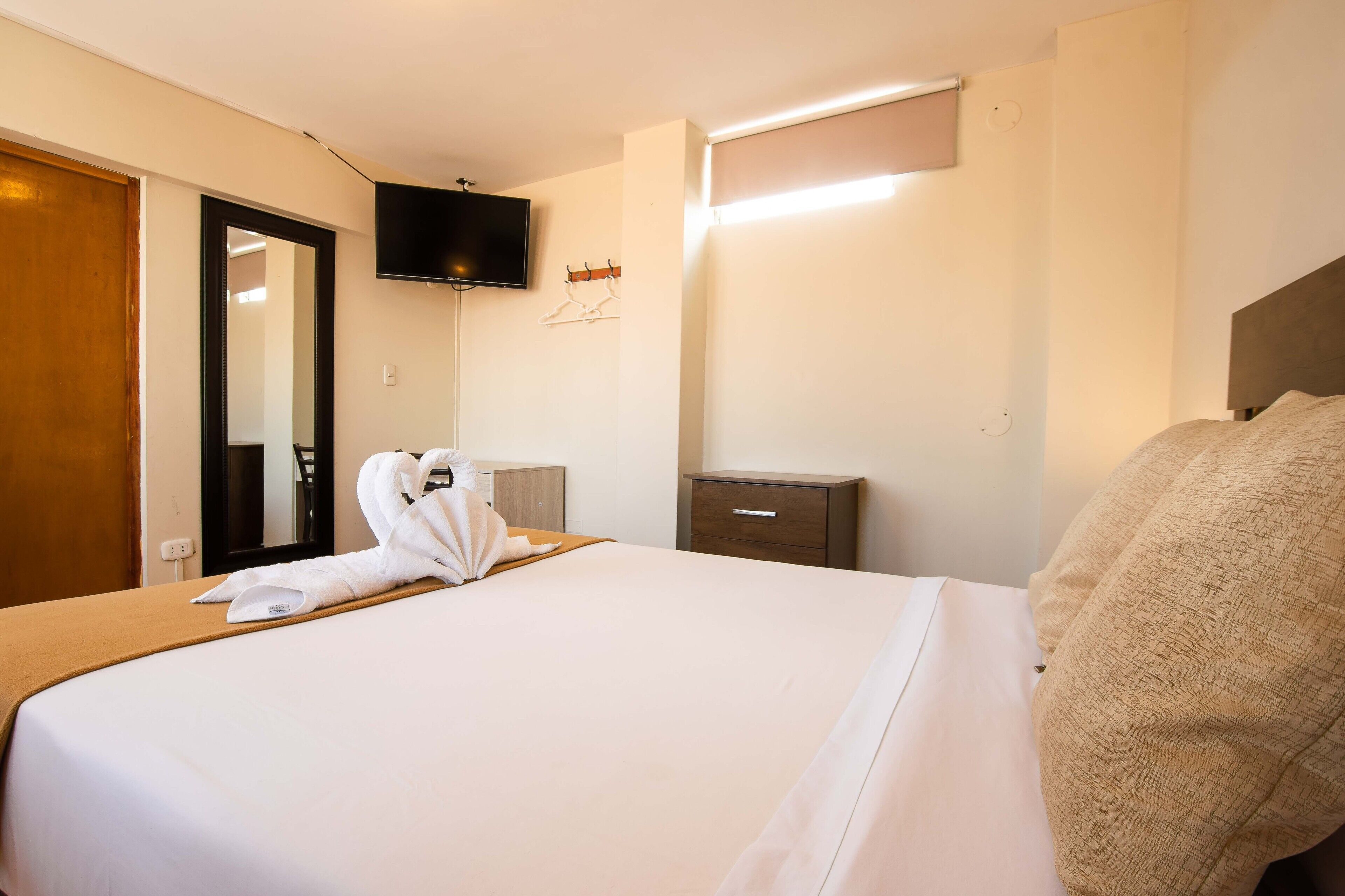 A hotel room with a neatly made bed, a flat-screen TV mounted on the wall, a wooden nightstand, and a wooden door.