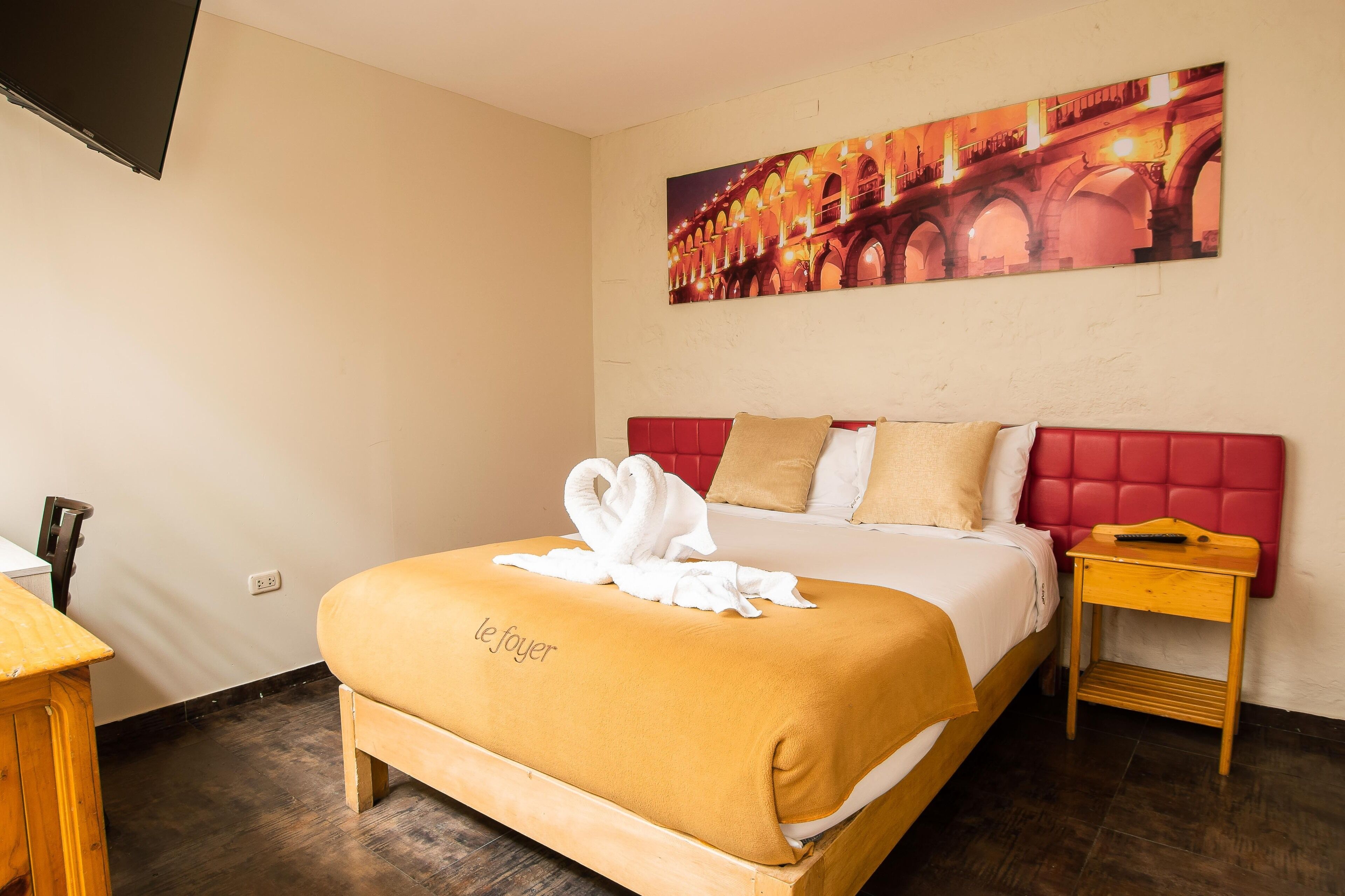 A hotel room with a bed, a red headboard, a wooden nightstand, a painting of a bridge, and a towel on the bed.