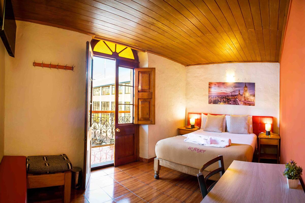 A bedroom with a bed, bedside tables, a desk, a chair, a bench, and a door leading to a balcony.