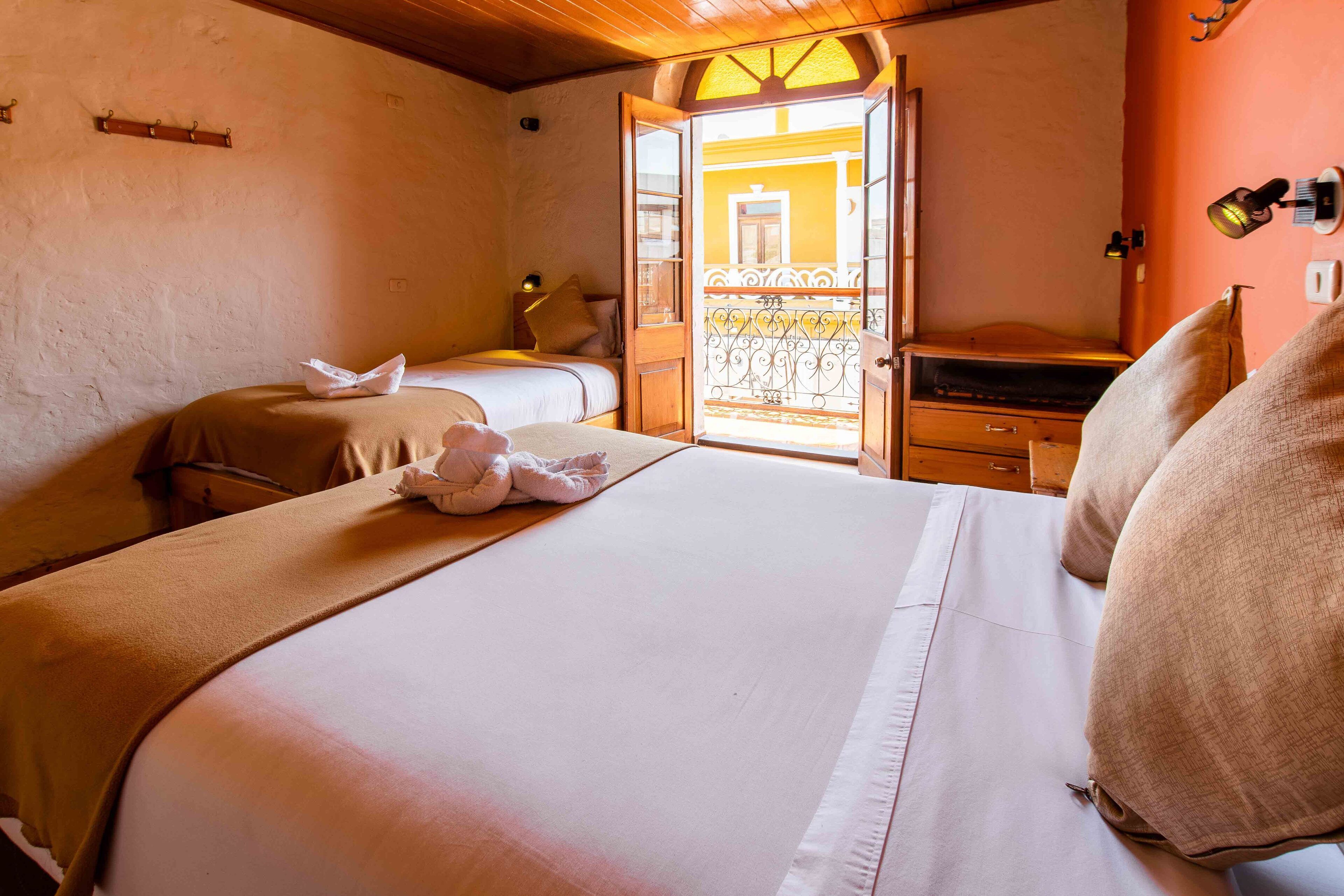 A hotel room with two beds, a wooden ceiling, a balcony with wrought iron railings, and a small desk with a lamp.