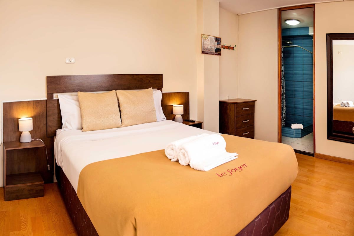 A hotel room with a bed, bedside lamps, a wooden nightstand, a wooden dresser, a mirror, and a shower area.