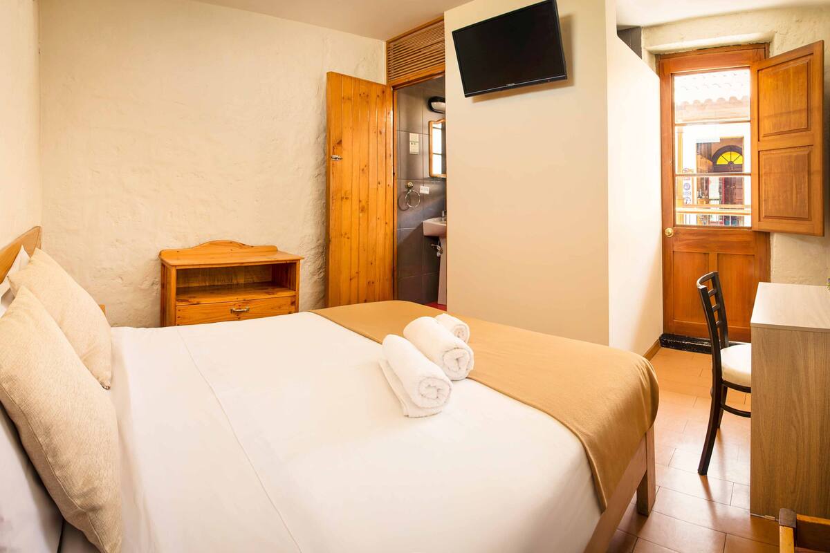 A hotel room with a bed, a wooden nightstand, a TV mounted on the wall, and a bathroom visible through an open door.