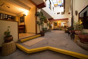A spacious indoor courtyard with a staircase leading to an upper level, surrounded by potted plants and wicker furniture. - Los Áticos (Cusco)