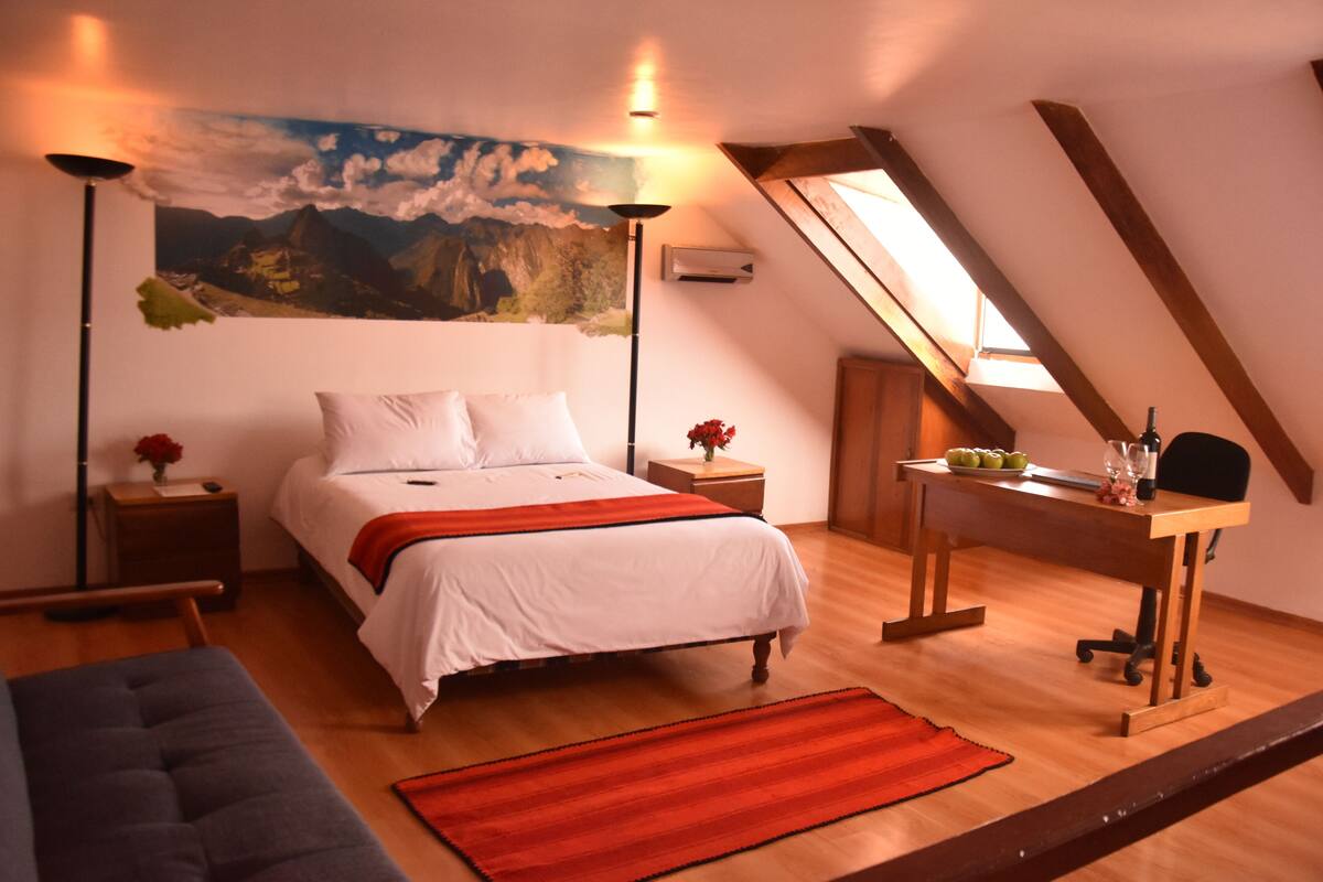 A bedroom with a bed, bedside tables, a desk, and a chair. There is a painting on the wall and a window with a view.