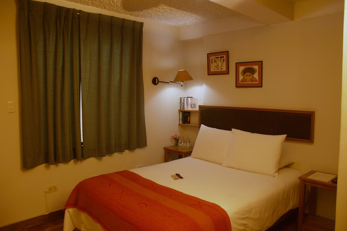 A hotel room with a bed, a nightstand, a lamp, a window with curtains, and two framed pictures on the wall.