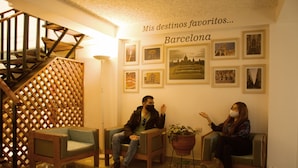 Un hall d’entrée avec deux fauteuils en bois, un escalier et un mur orné de tableaux et sur lequel on peut lire : « Mes destinations préférées… Barcelone ».