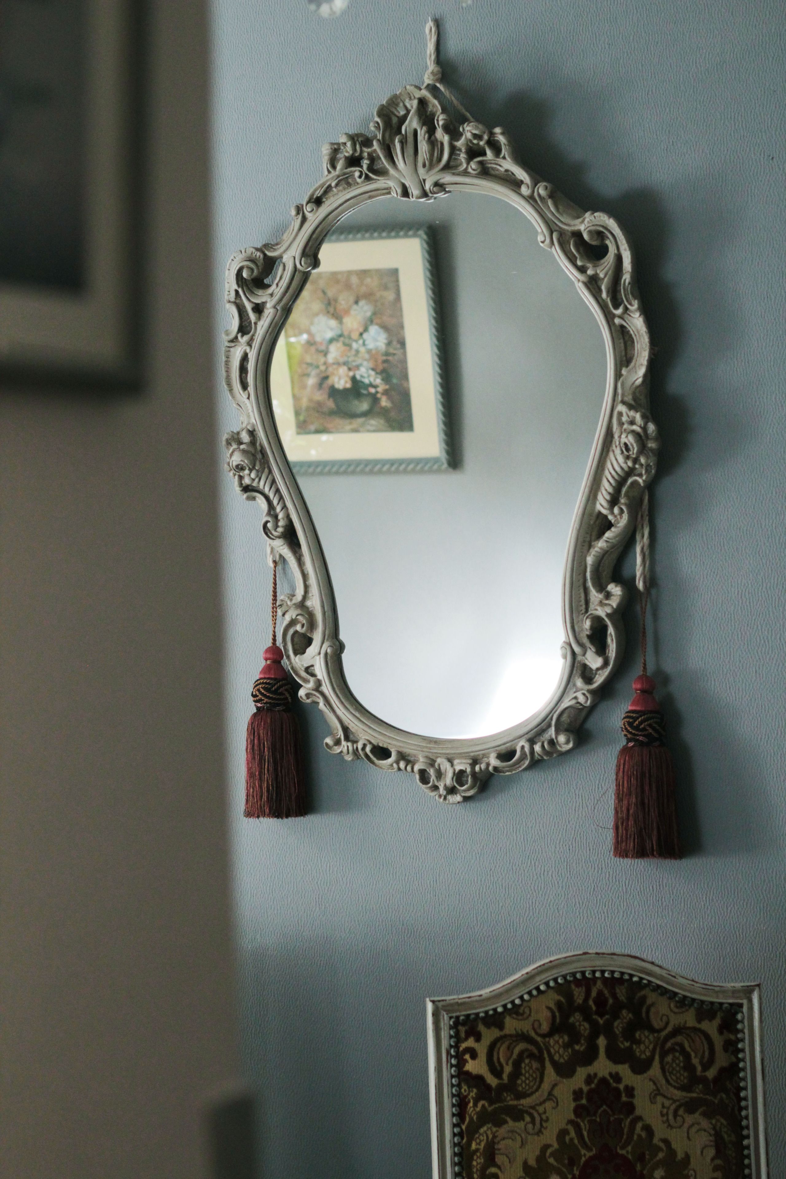 Um espelho com moldura ornamentada e franjas, fixado numa parede com um painel de cabeceira estampado.