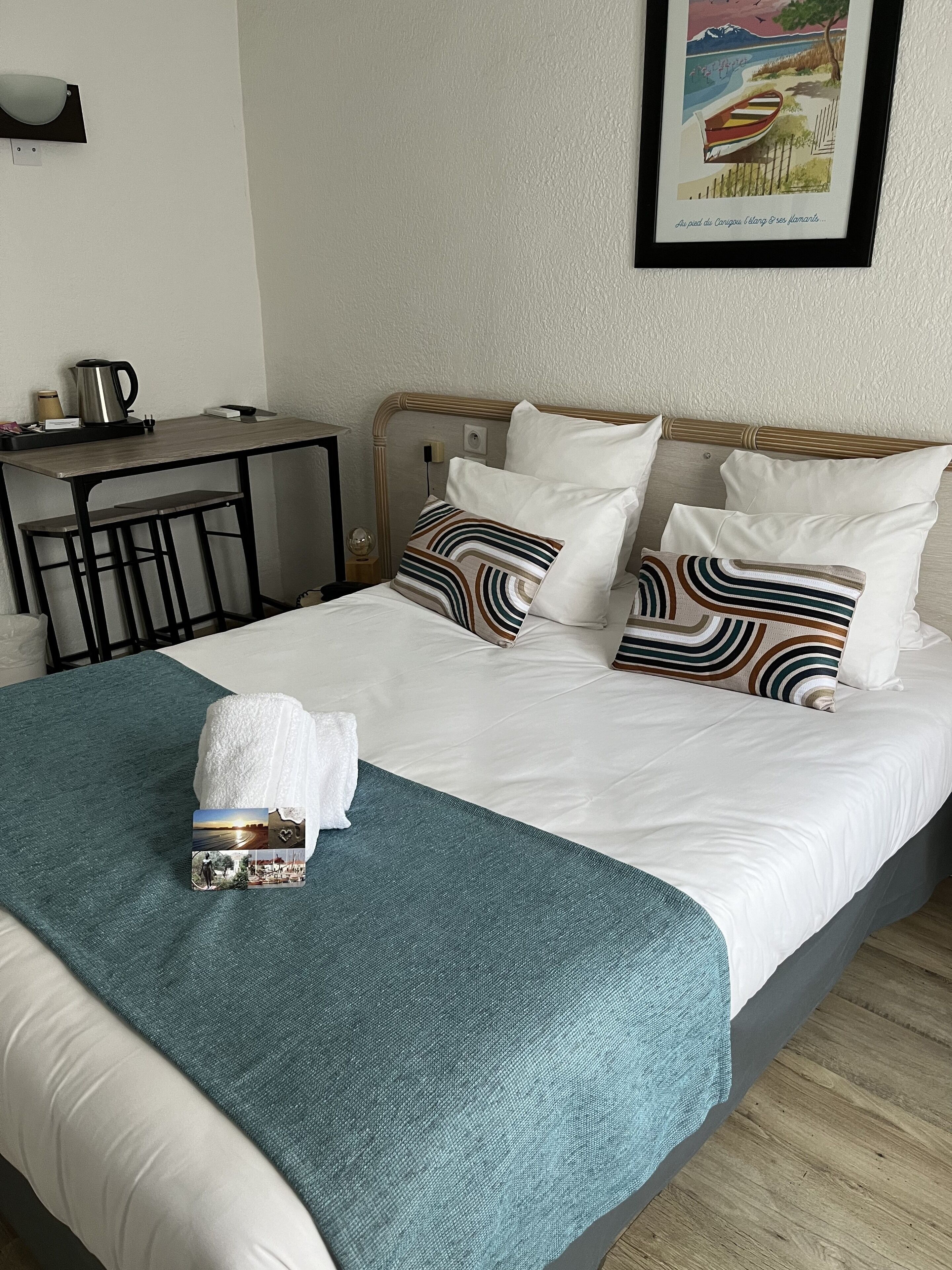 Un lit bien fait, agrémenté d’un plaid turquoise et de deux oreillers, une petite table avec une bouilloire et une tasse, et un tableau encadré au mur.