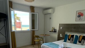 A hotel room with a bed, a sofa, a small table, a chair, a window with a view of the beach, and an air conditioning unit.