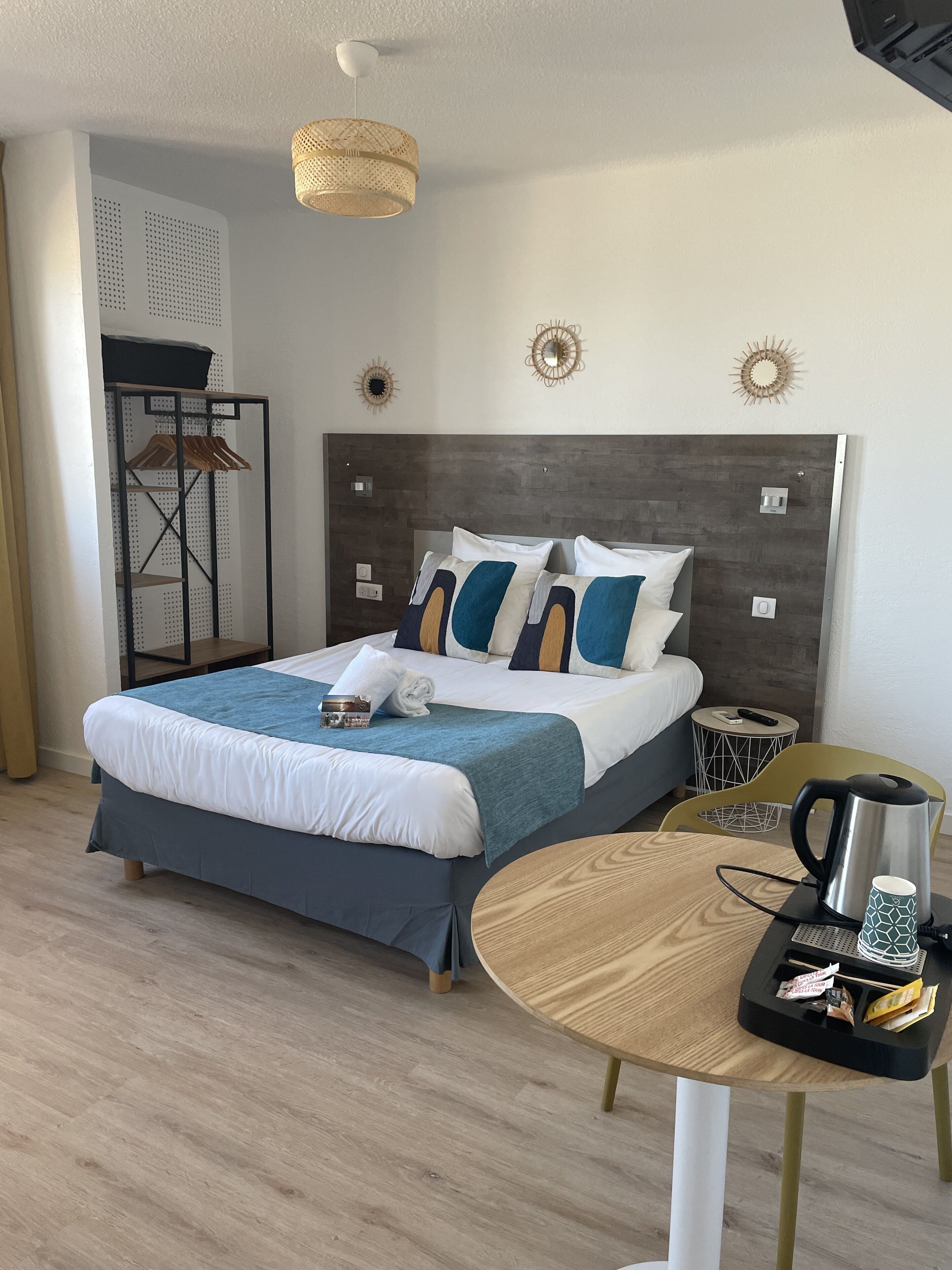 A modern hotel room with a neatly made bed, a round wooden table with a kettle and cups, and a small side table with a remote control.