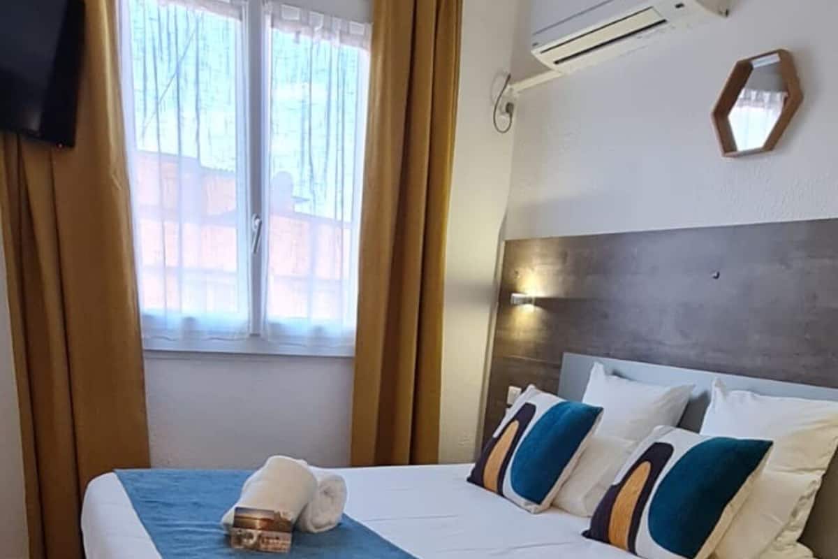A neatly made bed with a blue blanket and pillows, a flat-screen TV mounted on the wall, and a window with curtains.