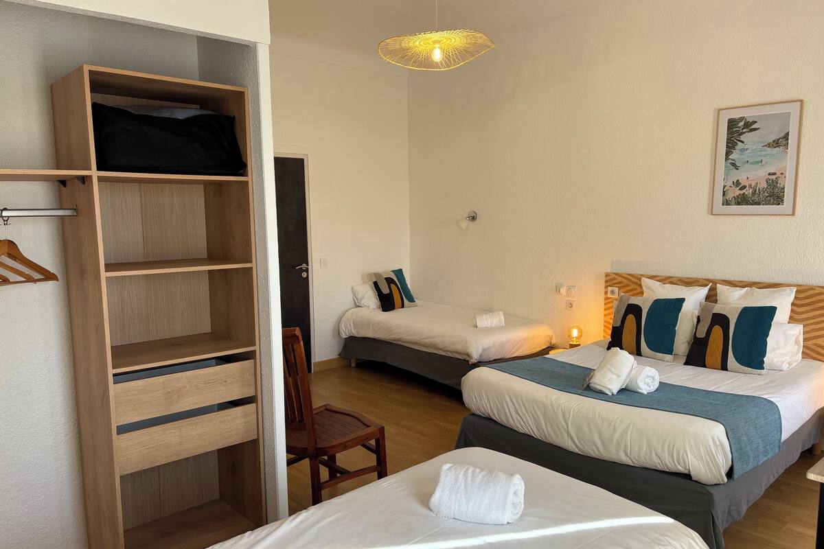 A hotel room with two beds, a wooden wardrobe, a chair, and a wall-mounted lamp.