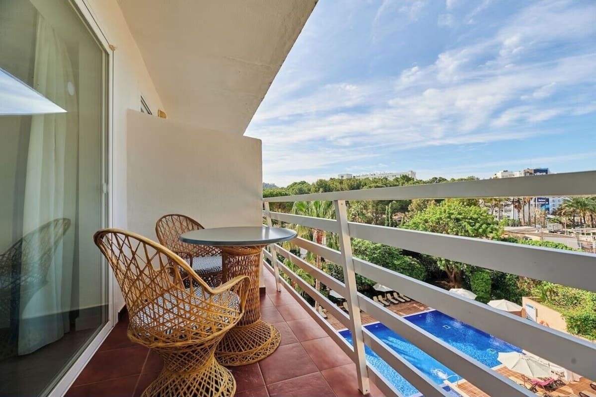 A balcony with wicker chairs and a small table, overlooking a swimming pool and greenery.