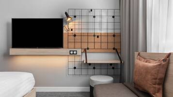 Une chambre d’hôtel moderne équipée d’un téléviseur à écran plat fixé au mur, d’une petite table de chevet, d’un banc rembourré et d’une étagère murale.