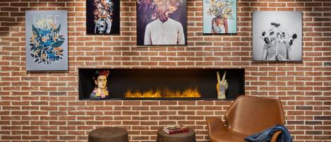 Una pared de ladrillo con una chimenea, cuatro cuadros enmarcados, un sillón de cuero marrón y dos otomanas.