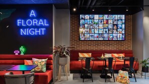 Un área de estar moderna con un letrero de neón que dice "A FLORAL NIGHT", una gran pantalla digital, asientos rojos y una pequeña mesa con sillas.