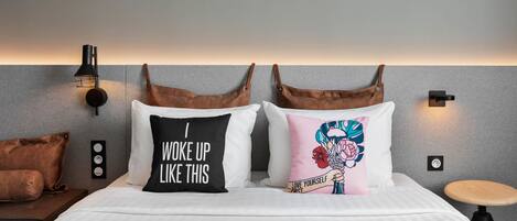 Una habitación de hotel con una cama, dos almohadas, una mesita de noche, una lámpara y una luz empotrada en la pared.