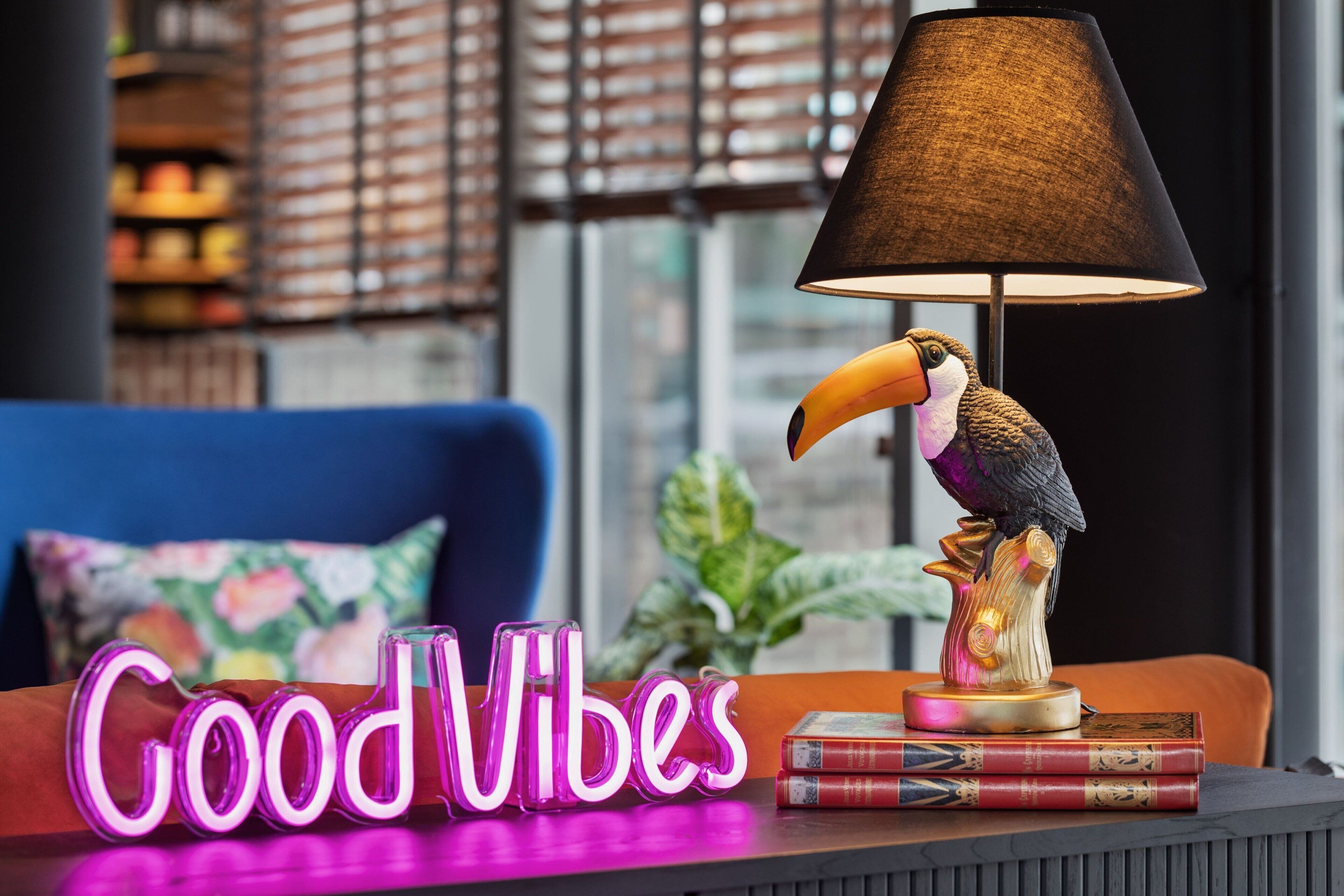 A room with a blue sofa, a lamp with a bird figurine, books, and a neon sign that reads "Good Vibes".