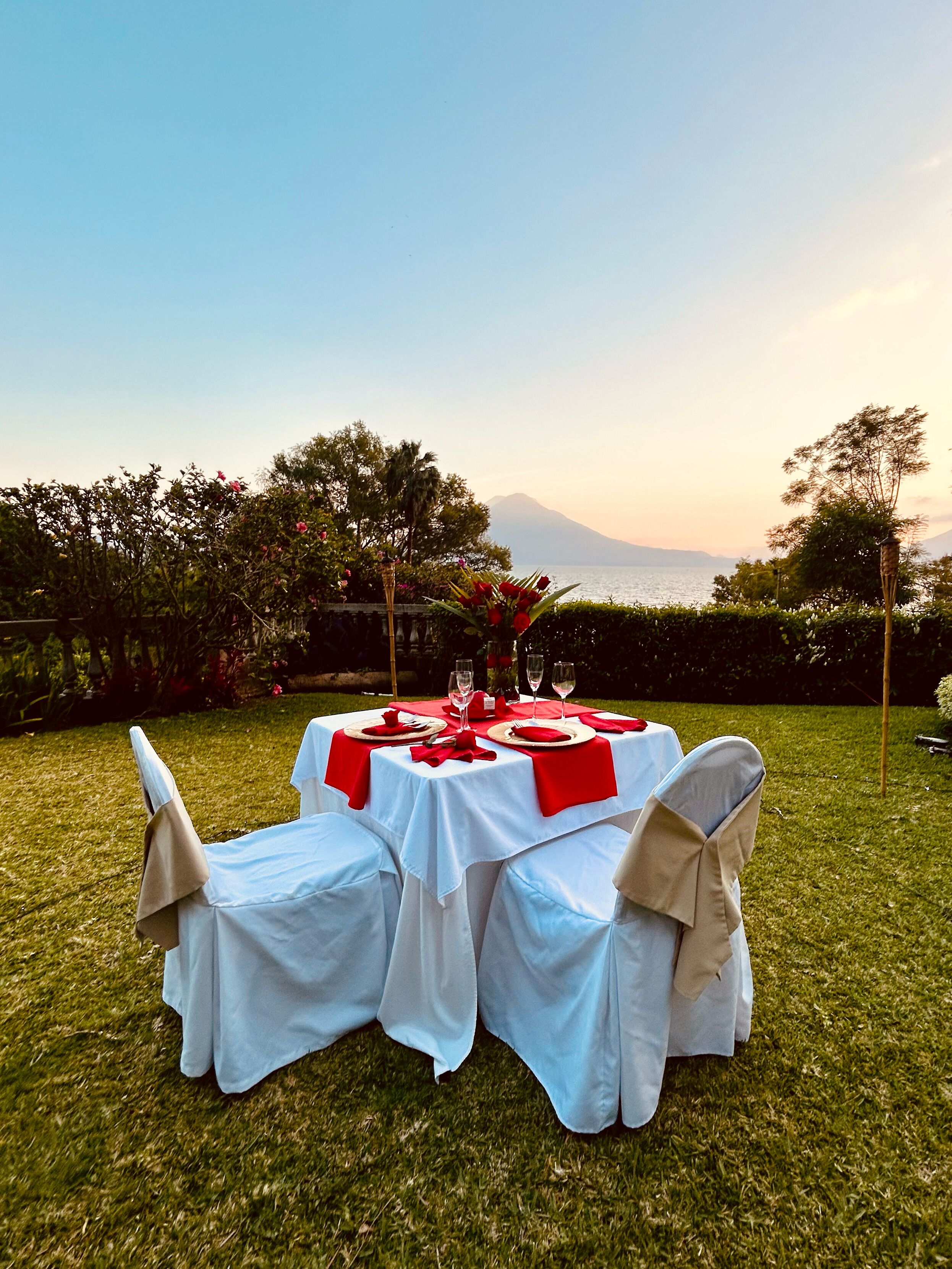 Mesa de comedor al aire libre preparada para cuatro personas, rodeada de follaje y con vistas al mar.