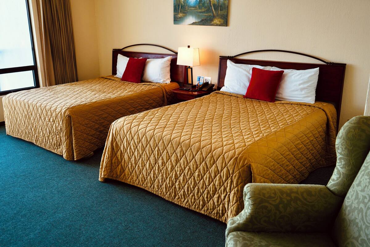 Habitación de hotel con dos camas, cada una con colcha acolchada y almohadas, una mesita de noche con lámpara y una silla.