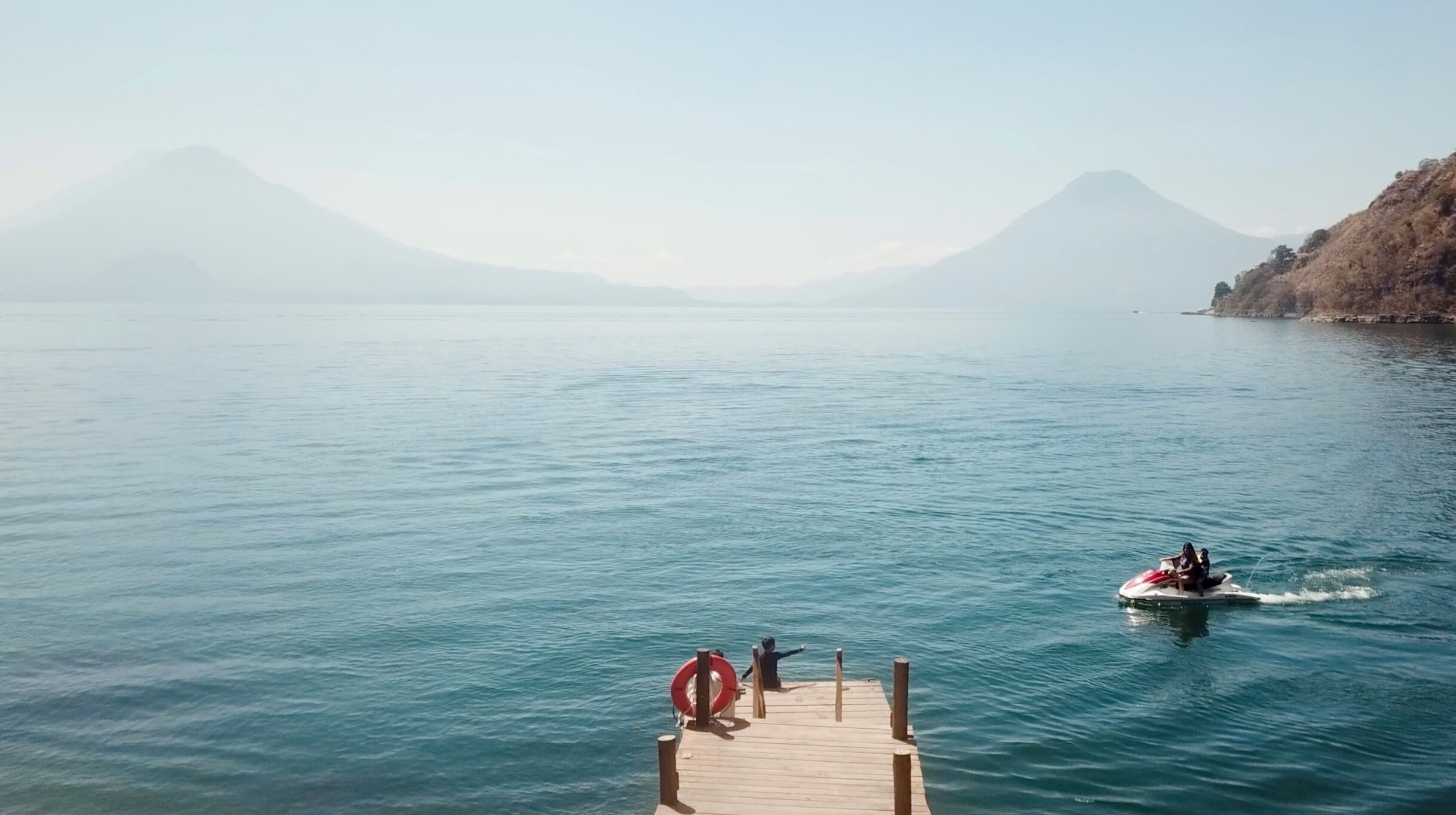 A calm lake with a wooden dock extending into the water, a boat moving away, and mountains in the background.