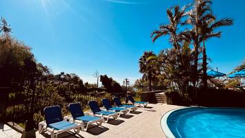 Zona junto a la piscina con sillas reclinables, cielo azul despejado y palmeras.