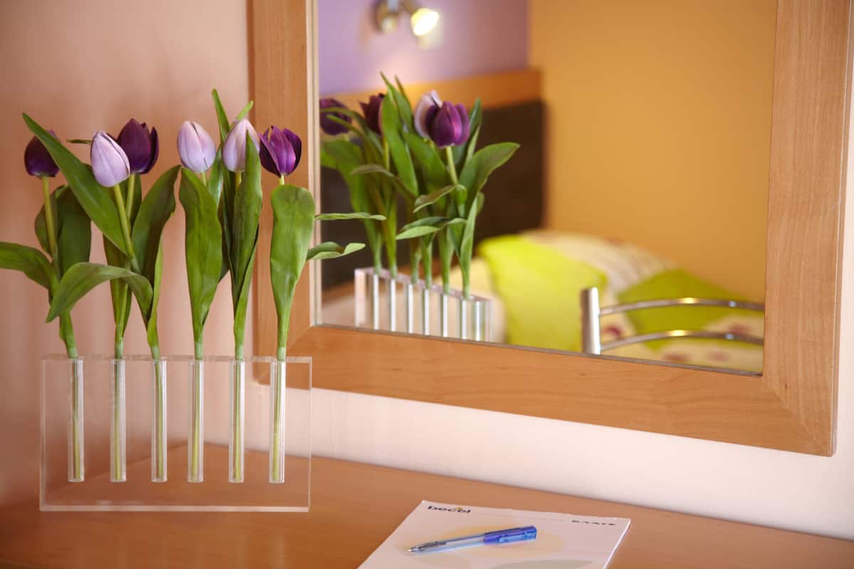Un escritorio de madera con un florero transparente que contiene tulipanes morados, un espejo que refleja la habitación y un bolígrafo sobre un papel.