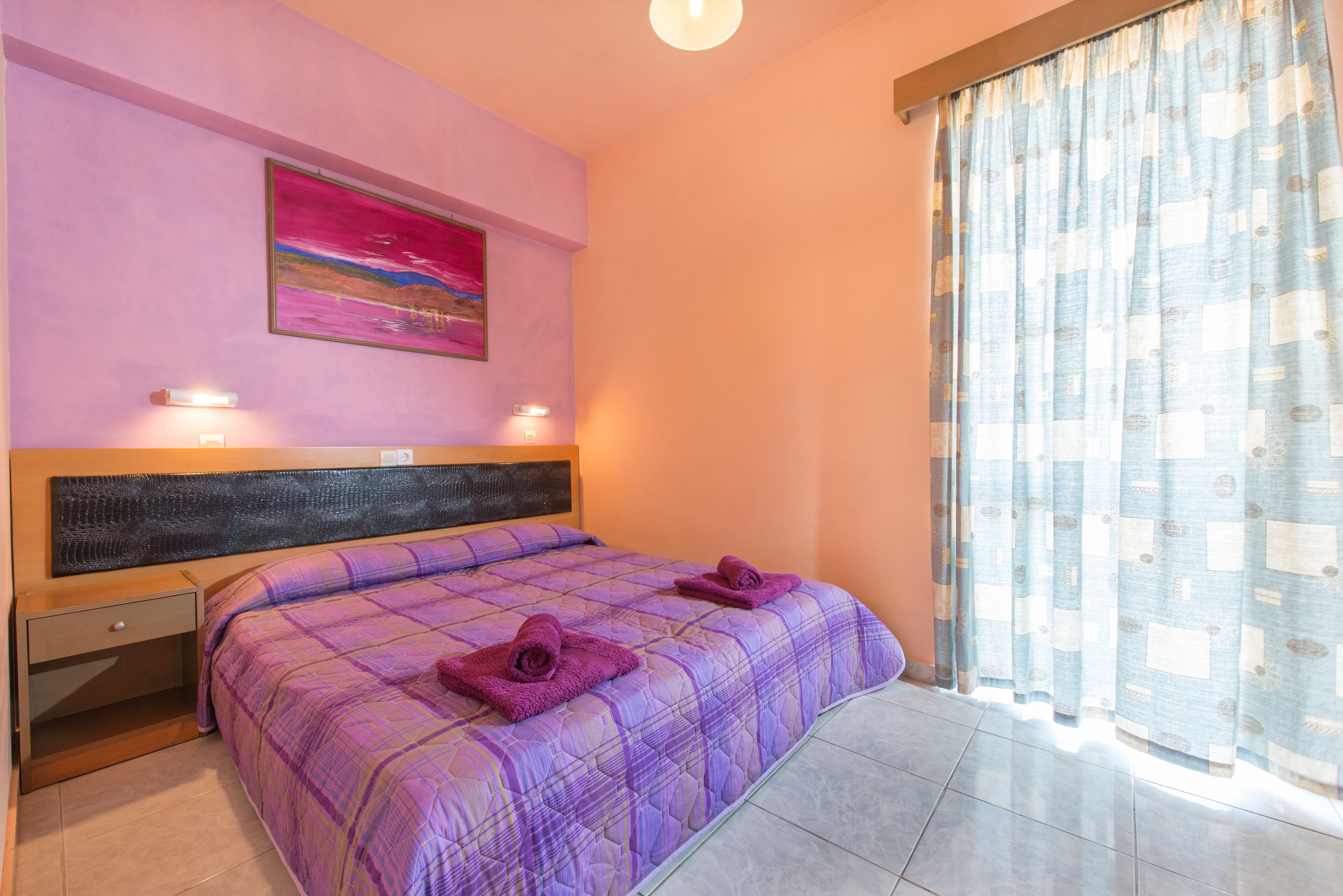 A bedroom with a bed, a nightstand, a wall-mounted artwork, and a window with curtains.