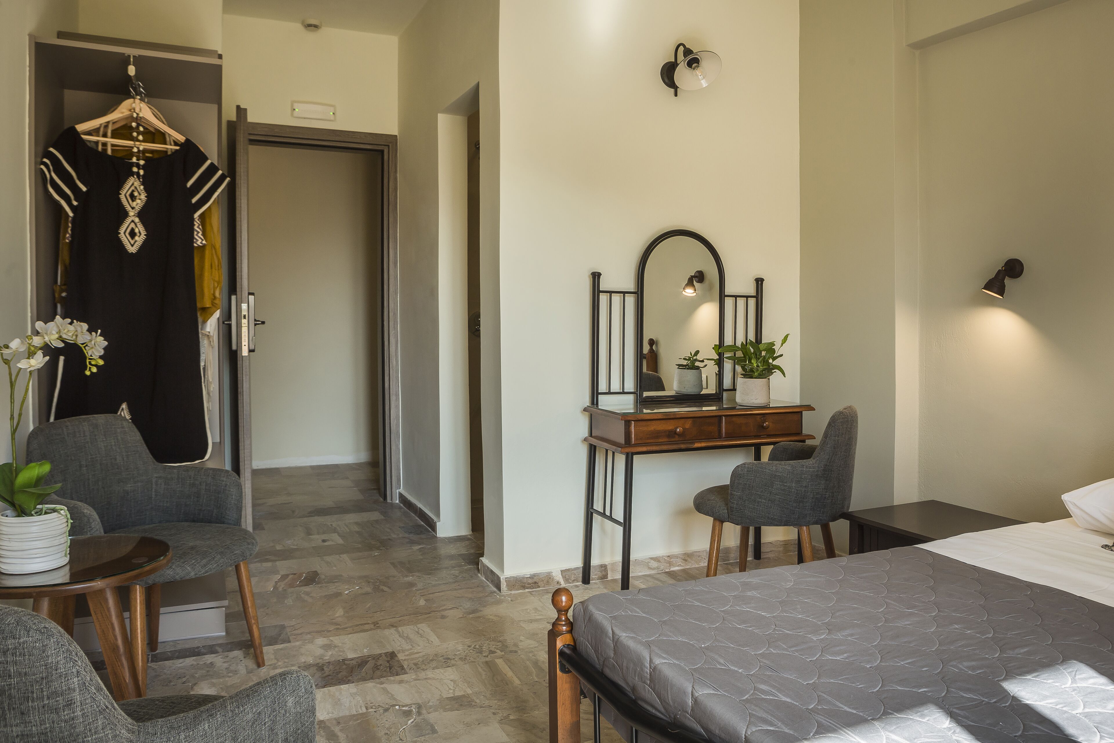 A modern hotel room with a bed, a desk, a chair, a hanging garment, and a potted plant.