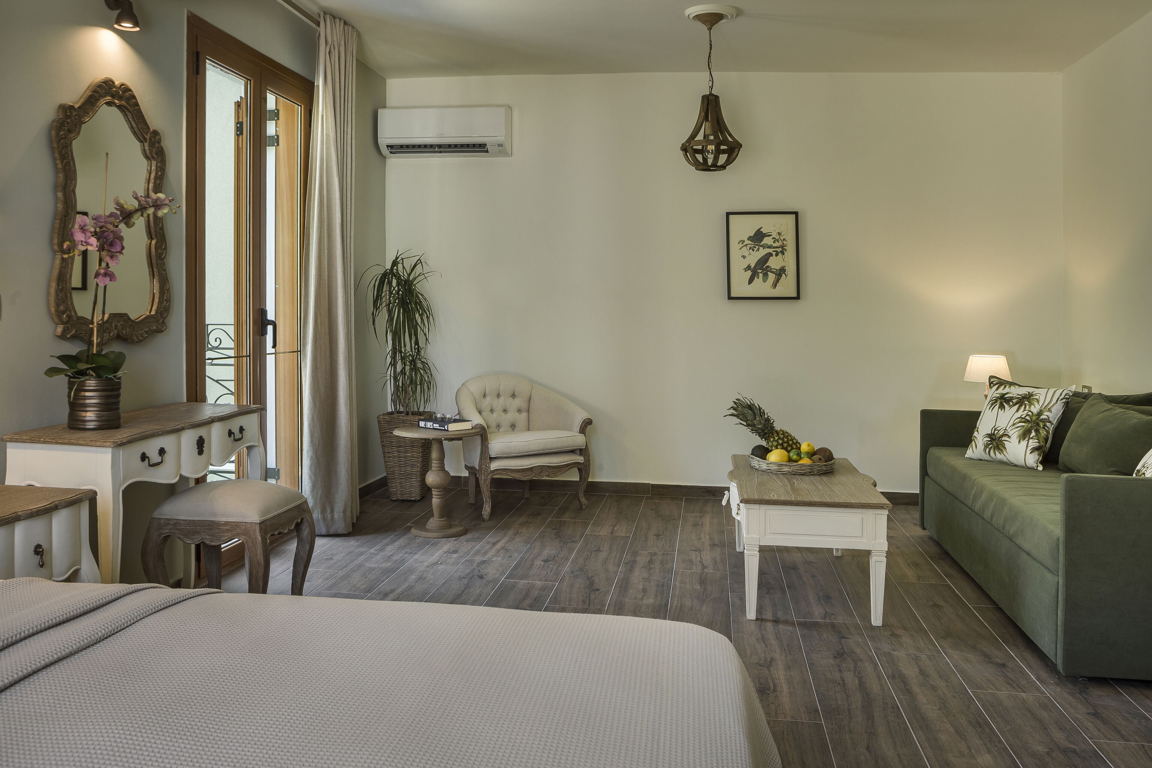 A hotel room with a bed, a desk, a chair, a sofa, a small table with fruit, a hanging light fixture, and a wall-mounted air conditioner.