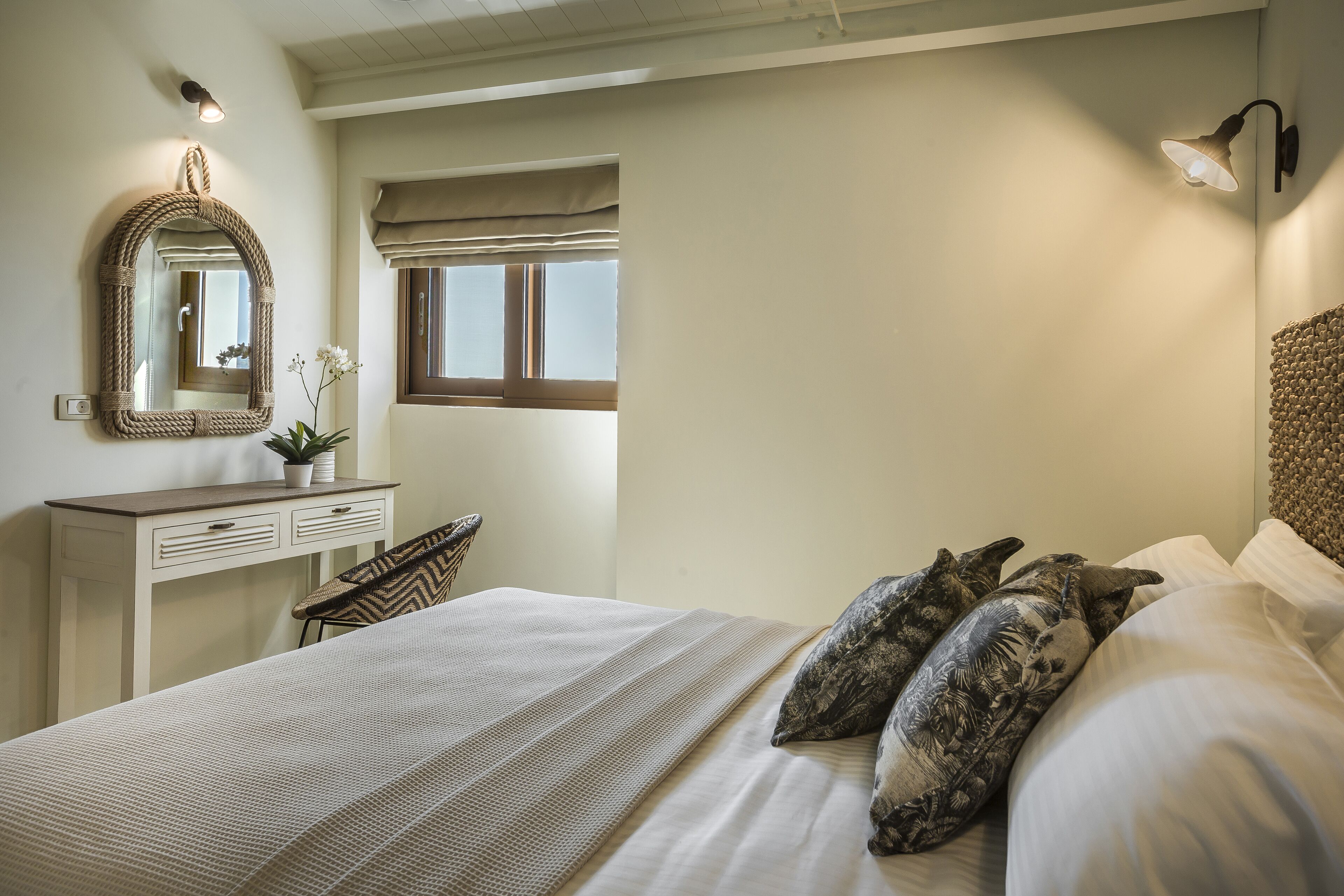 A bedroom with a bed, a bedside table, a mirror, a window with blinds, and a wall-mounted lamp.
