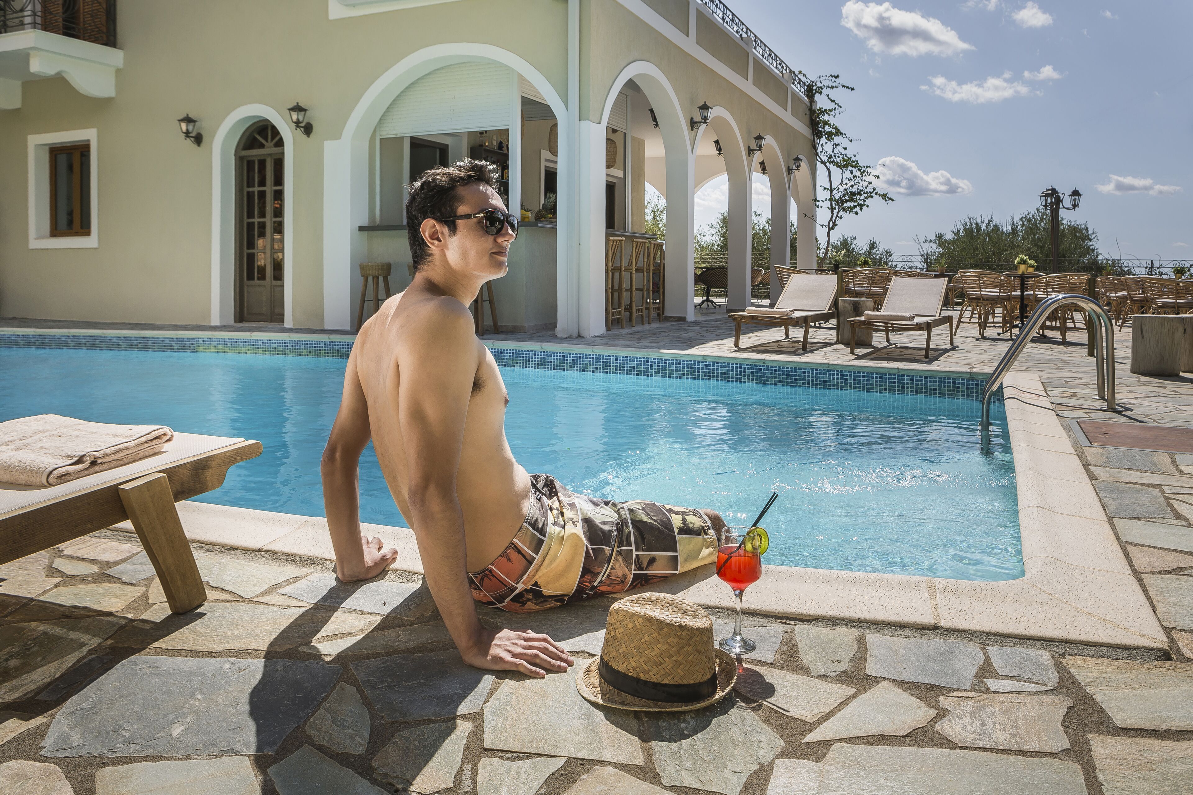 A pool area with a person sitting by the pool, a drink, and a straw hat.