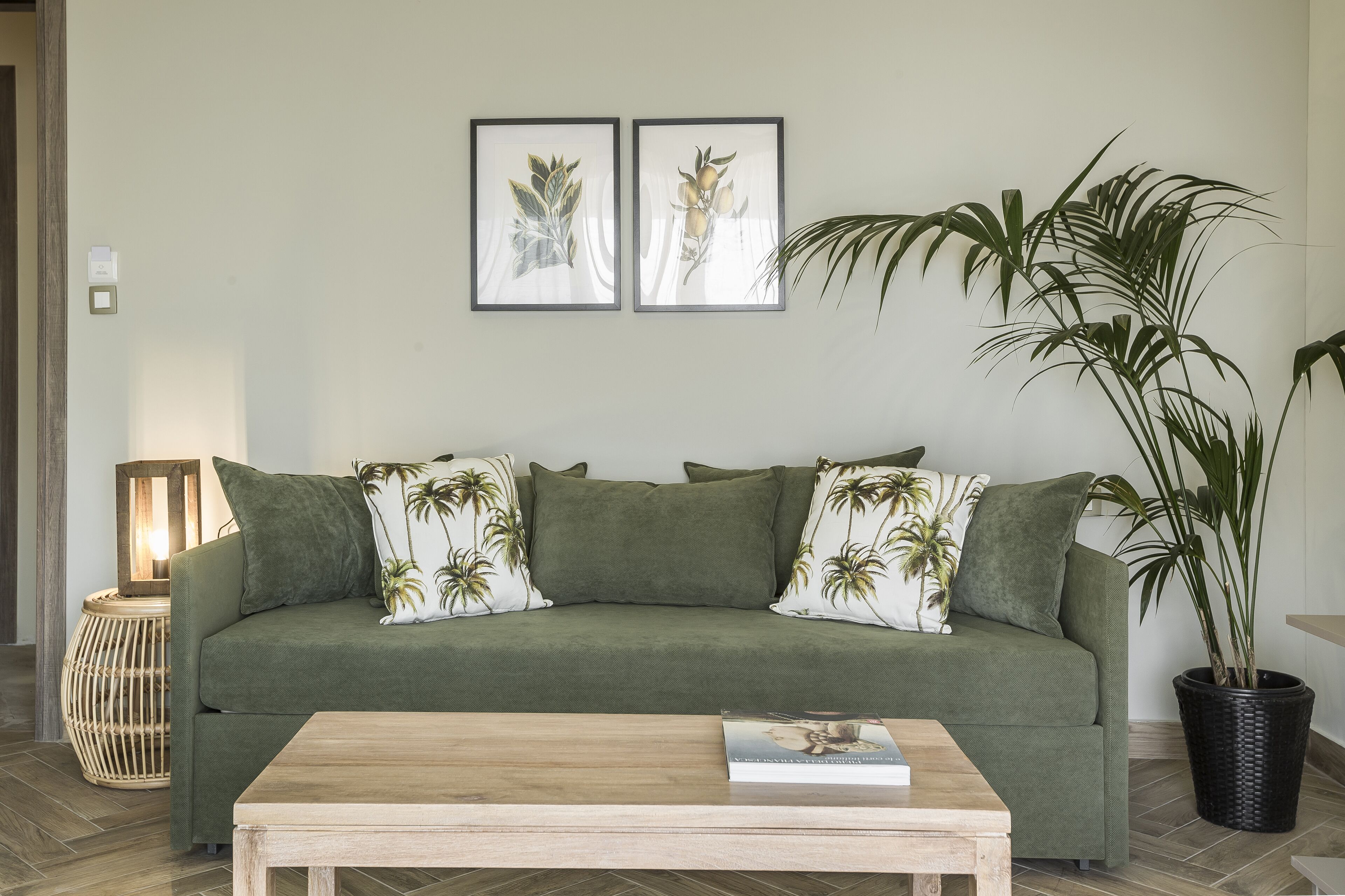 Un sofá verde con cojines estampados de hojas de palmera, una mesa de centro de madera y dos láminas botánicas enmarcadas en la pared.