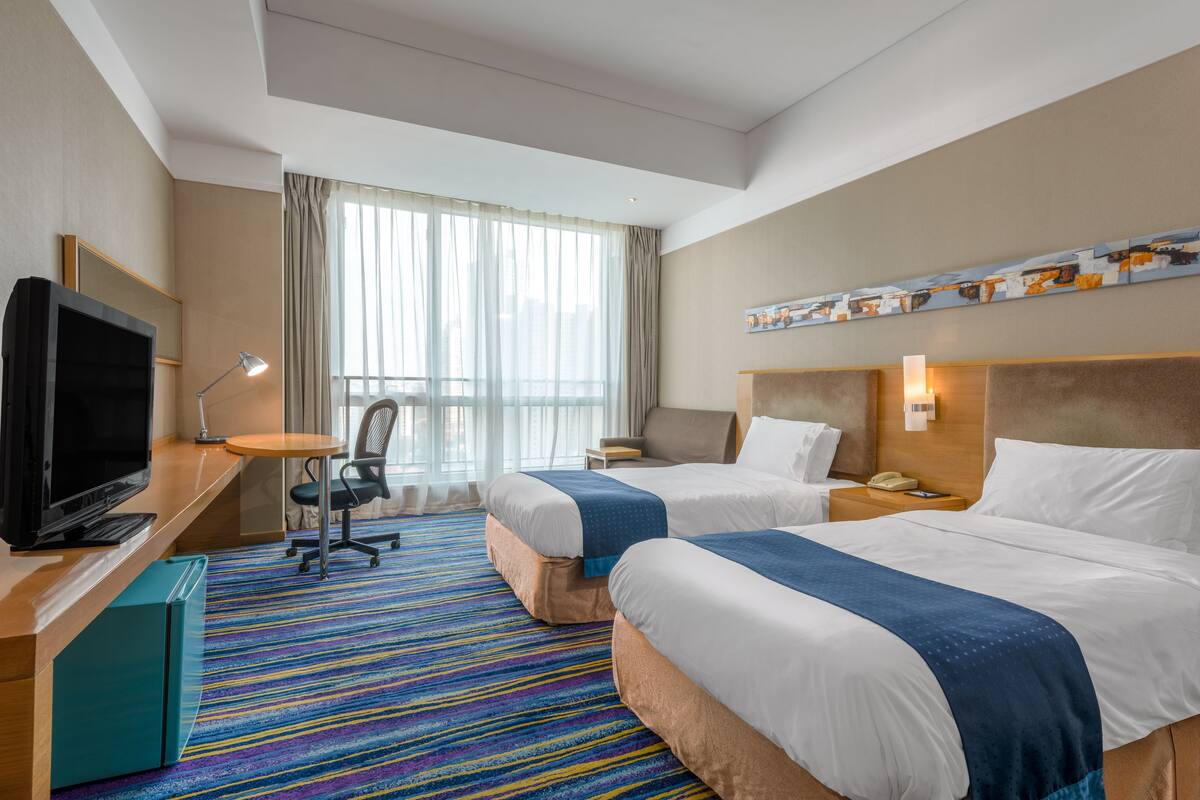 A hotel room with two beds, a desk with a TV, a chair, a small table, and a colorful striped carpet.