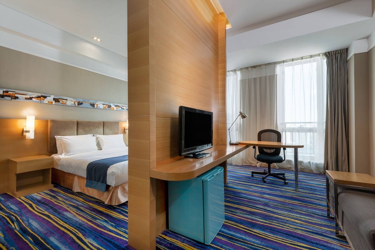 A hotel room with a bed, a desk with a television, a chair, a small refrigerator, and a window with curtains.