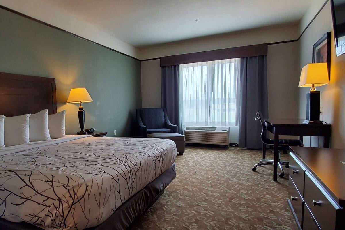 A hotel room with a large bed, two bedside lamps, a desk with a chair, a window with curtains, and an air conditioning unit.