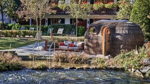 A rustic wooden structure with a door, a small outdoor seating area with a sofa and chairs, and a view of a river. - Spa & Resort Bachmair Weissach, LUXURY FAMILY RESORT (Kreuth)