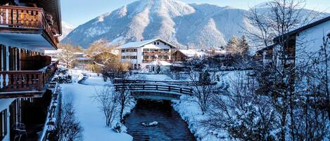 A snow-covered mountain village with a wooden bridge over a stream, traditional buildings, and a clear view of the mountains.