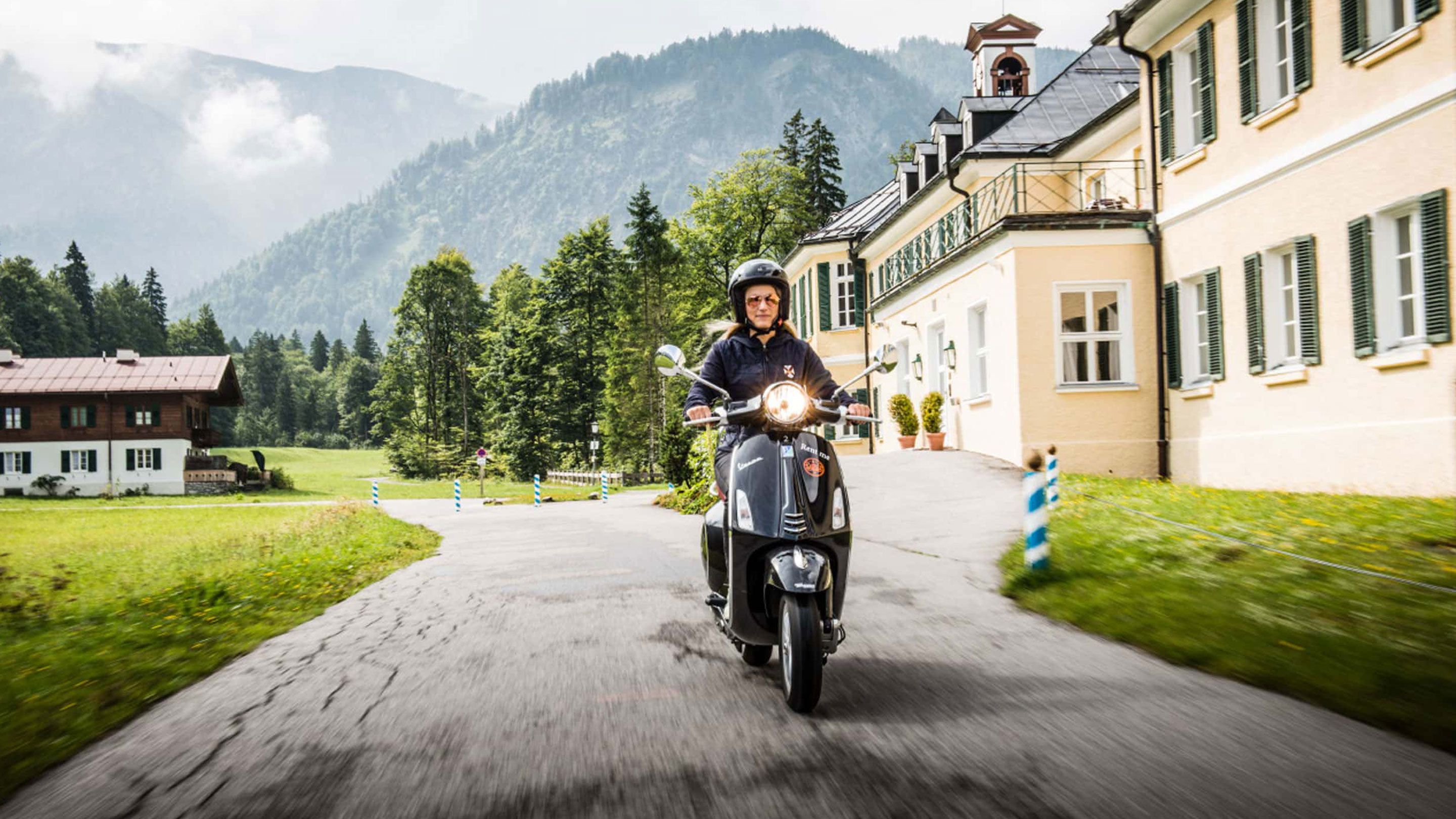A person riding a scooter on a paved road with a building and trees in the background.