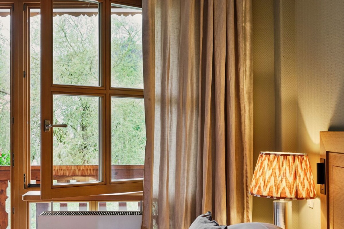 A bedroom with a bed, a window with curtains, a lamp, and a view of greenery outside.