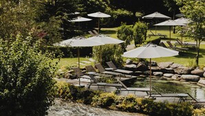 A serene outdoor setting with a pond, lounge chairs, and umbrellas. - Spa & Resort Bachmair Weissach, LUXURY FAMILY RESORT (Kreuth)
