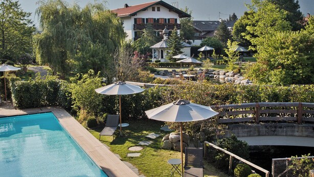 A swimming pool with a wooden deck, lounge chairs, umbrellas, and a view of mountains. - Spa & Resort Bachmair Weissach, LUXURY FAMILY RESORT (Kreuth)